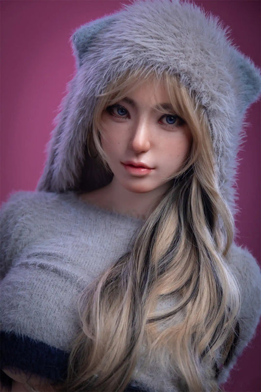 Angelina sex doll, long blonde highlighted wavy hair with curtain fringe, deep-set blue eyes and parted lips with natural flush, grey fuzzy bear-ear crop hoodie hugging full bust, three-quarter portrait shot with arms crossed in self-embrace, warm rose-pink background – lifelike Russian chubby curvy sex doll by TopLoveDoll